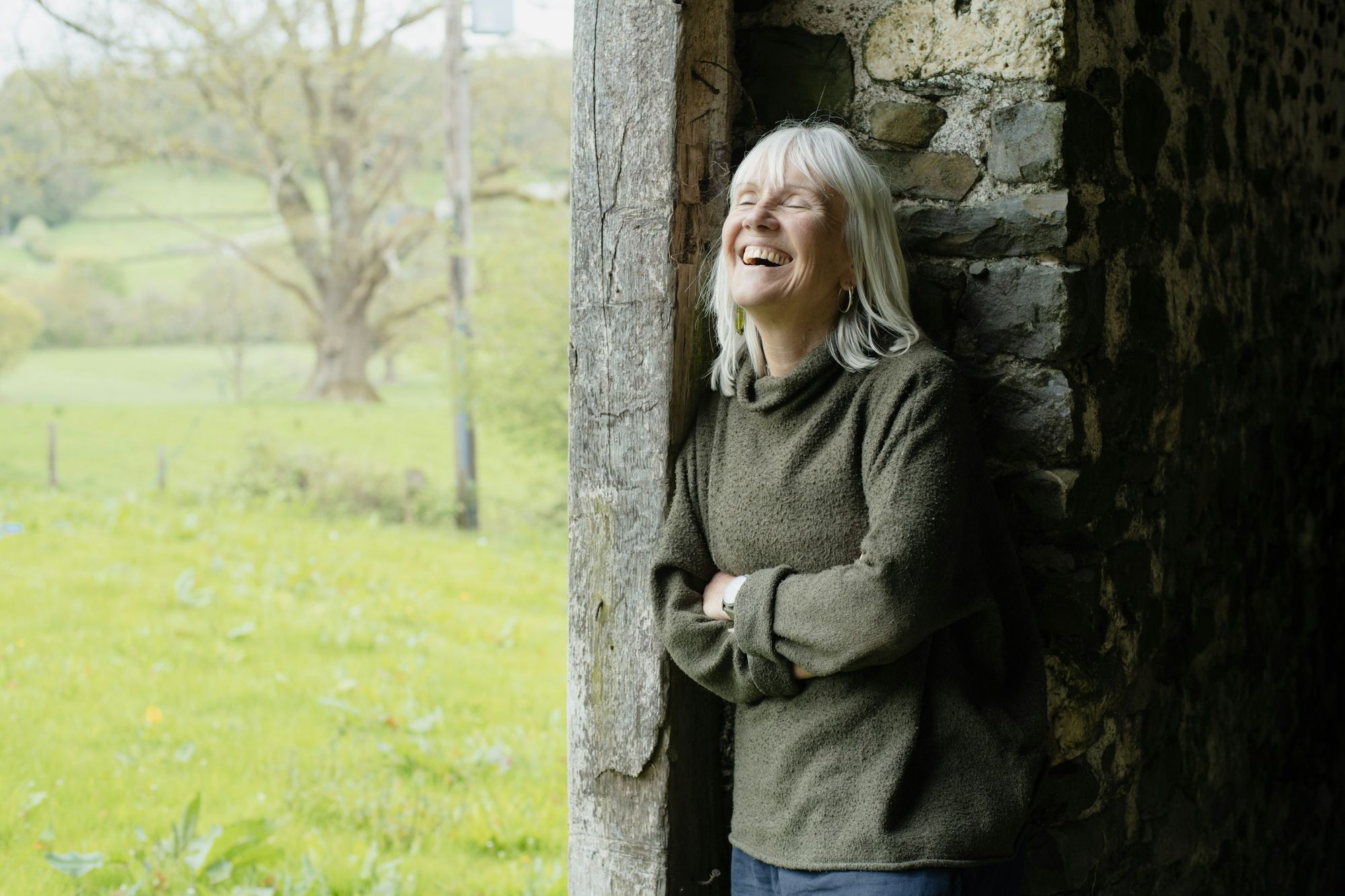 A woman laughing in the doorway of a stone barn, green fields behind her